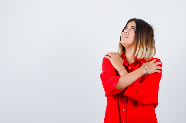 Mooie vrouw die zichzelf omhelst, opkijkt in een rode blouse en zich afvraagt. vooraanzicht.