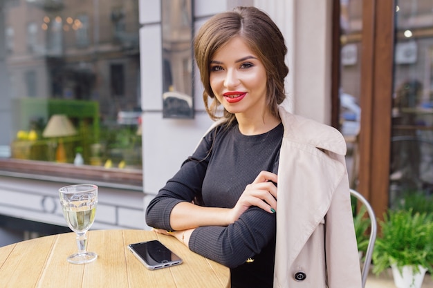 Mooie vrouw gekleed in zwarte jurk en beige loopgraaf met stijlvol kapsel en rode lippen op een terras