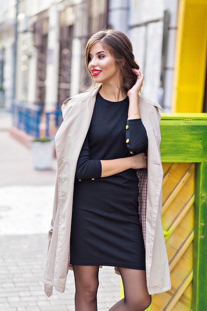 Mooie vrouw gekleed in zwarte jurk en beige loopgraaf met stijlvol kapsel en rode lippen op straat