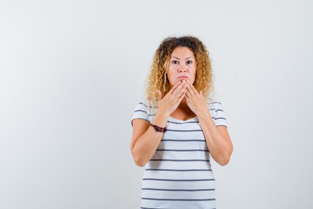 Mooie vrouw hand in hand op kin in t-shirt en kijkt verrast, vooraanzicht.