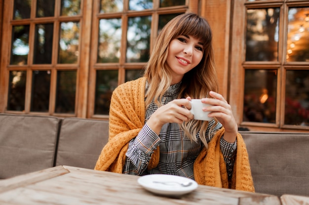 Gratis foto mooie vrouw hebben een koffiepauze in gezellig café met houten interieur, praten via de mobiele telefoon. holding kopje warme cappuccino. winter seizoen. het dragen van een elegante jurk en een gele plaid.