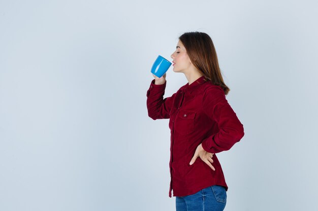 Mooie vrouw in bordeauxrode blouse die thee drinkt terwijl ze de hand op de taille houdt en er dorstig uitziet.