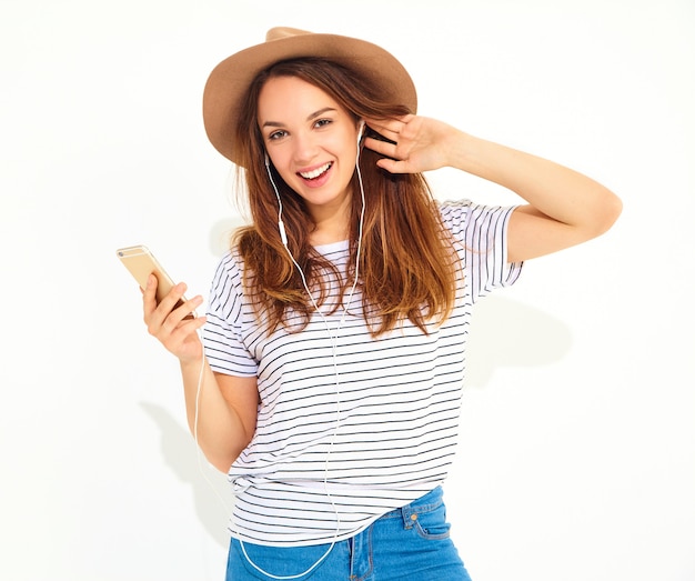 mooie vrouw in casual hipster zomer kleding zonder make-up in bruine hoed luisteren muziek in telefoon met koptelefoon op wit wordt geïsoleerd