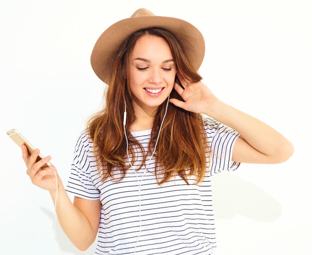 mooie vrouw in casual hipster zomer kleding zonder make-up in bruine hoed luisteren muziek in telefoon met koptelefoon op wit wordt geïsoleerd