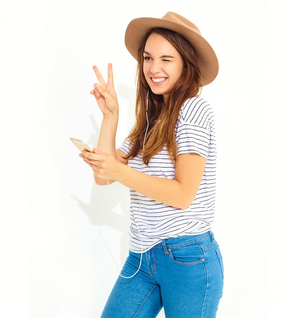 mooie vrouw in casual hipster zomer kleding zonder make-up in bruine hoed luisteren muziek in telefoon met koptelefoon op wit wordt geïsoleerd