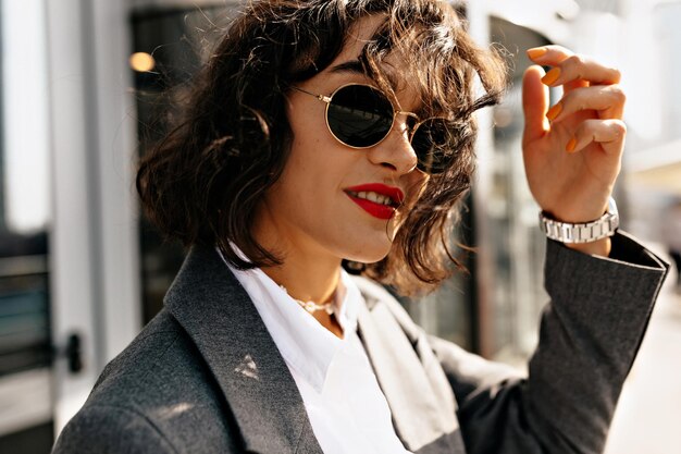 Mooie vrouw in grijze jas en wit overhemd in zonnebril met rode lippenstift raakt haar aan en poseert in de buurt van haar huis Mooie dame in grijze oversized jas en broek loopt naar buiten