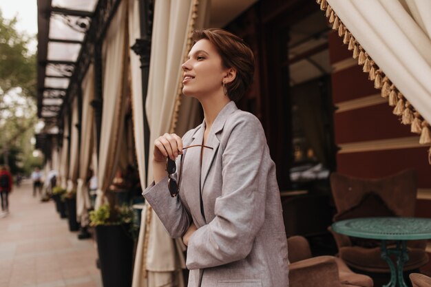Mooie vrouw in grijze outfit poseren in de buurt van straatcafé. Kortharig charmant meisje in oversized jas glimlacht en geniet buiten van de lentedag