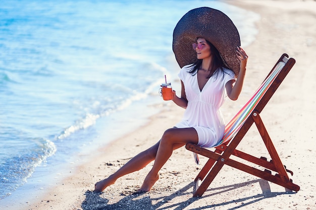 Mooie vrouw in grote strohoed ontspannen op een ligstoel strand en drinkt verfrissend sap zomer vakantie...