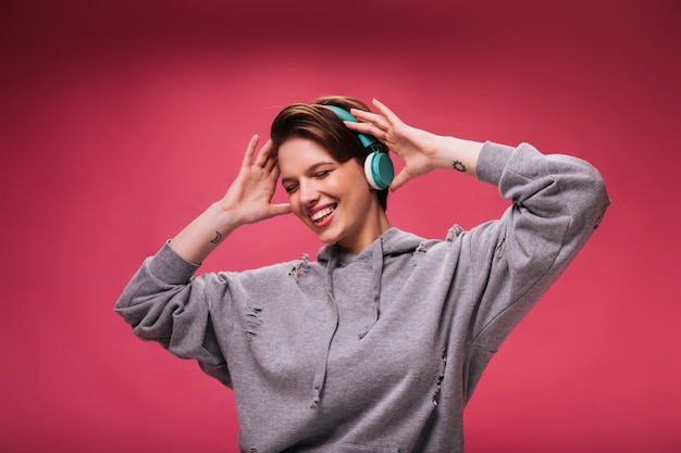 Mooie vrouw in hoodie luisteren naar muziek in koptelefoon op roze achtergrond. Tienermeisje in grijs sweatshirt dansen en glimlachen op geïsoleerd