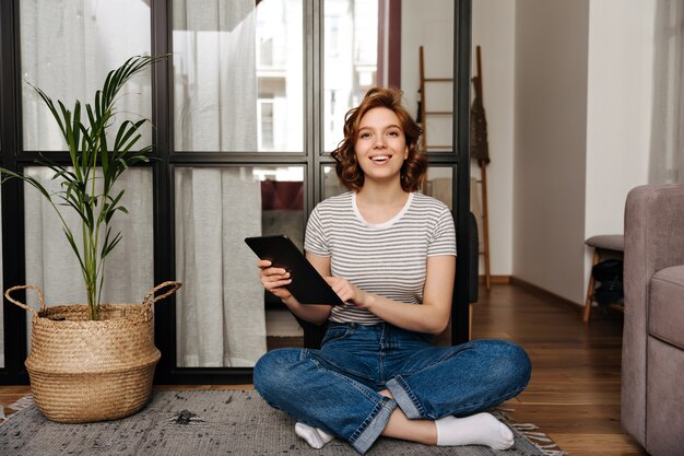 Mooie vrouw in jeans broek zit op de vloer, houdt tablet en kijkt naar de camera.