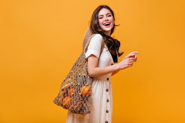 Mooie vrouw in katoenen jurk lacht en houdt string tas met fruit. Portret van dame met golvend haar op oranje achtergrond.