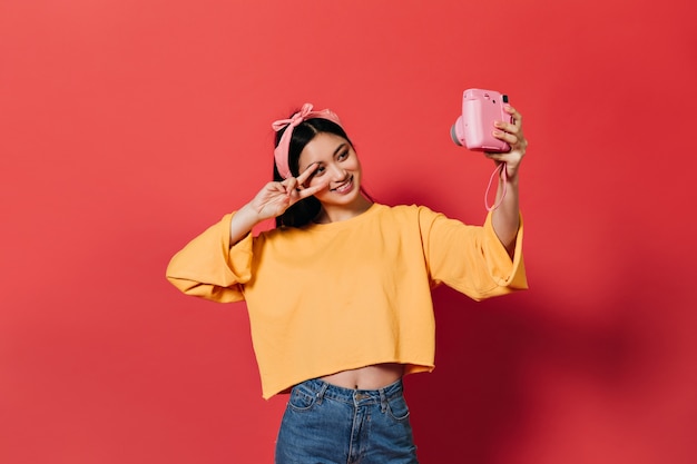 Mooie vrouw in oranje trui en spijkerbroek vertoont vredesteken en maakt selfie