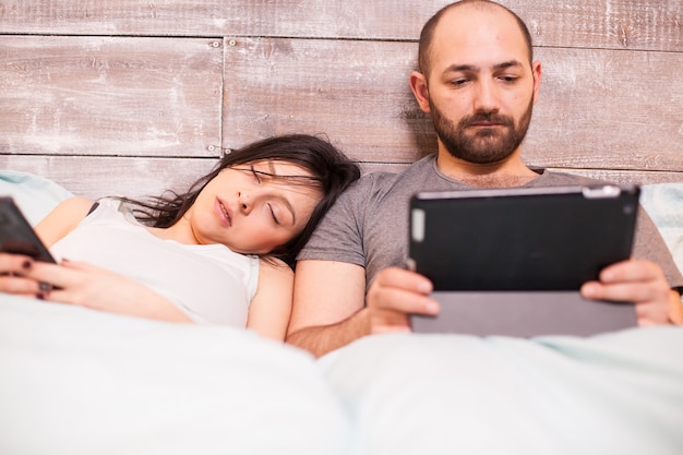 Mooie vrouw in pyjama slapen terwijl haar man aan tabletcomputer werkt.