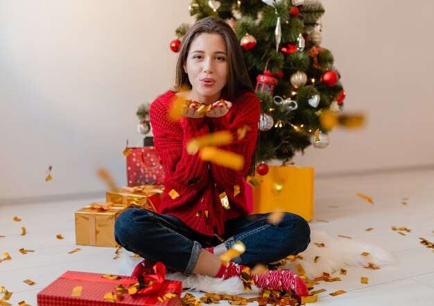 Mooie vrouw in rode trui om thuis te zitten op kerstboom gouden confetti gooien omringd met cadeautjes en geschenkdozen