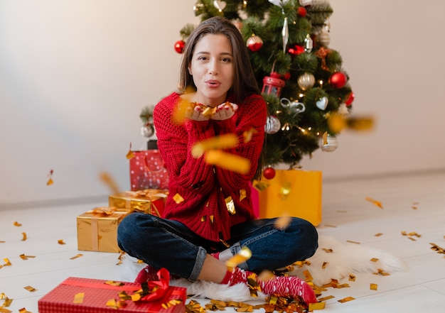 Mooie vrouw in rode trui om thuis te zitten op kerstboom gouden confetti gooien omringd met cadeautjes en geschenkdozen