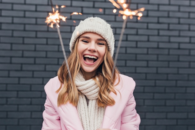 Mooie vrouw in winteraccessoires positieve emoties uitdrukken en wonderkaarsen zwaaien. Buiten foto van prachtig meisje in roze hoed Nieuwjaar vieren.