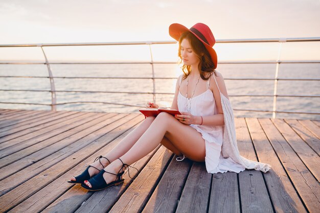 Mooie vrouw in witte jurk zittend aan zee op zonsopgang denken en maken van aantekeningen in dagboekboek