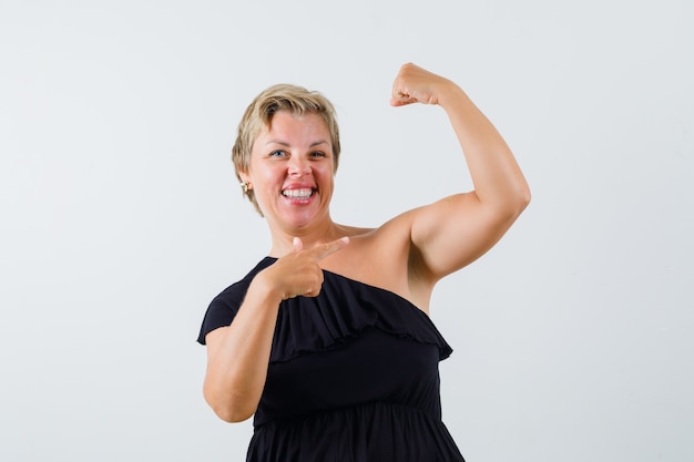 Mooie vrouw in zwarte blouse wijzend op haar armspieren en zelfverzekerd op zoek