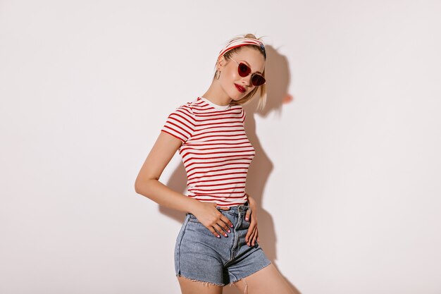 Mooie vrouw met blond kapsel en rode lippenstift in zonnebril, gestreept stijlvol t-shirt en denim shorts glimlachend op geïsoleerde muur