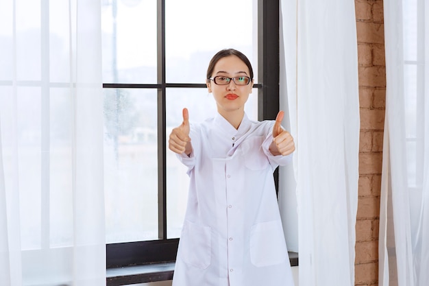 Mooie vrouw met bril in laboratoriumjas permanent in de buurt van raam en duimen opdagen.