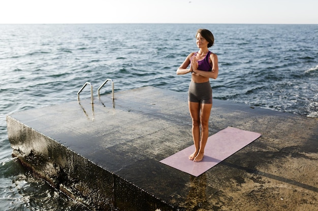 Mooie vrouw met donker kort haar dat met gesloten ogen staat en mediteert aan zee. mooie dame in sportieve top en korte broek die yoga doet met uitzicht op zee op de achtergrond Gratis Foto