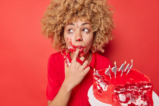 Gratis foto mooie vrouw met doordachte uitdrukking likt vinger besmeurd met zoete room eet heerlijke cake ergens geconcentreerd draagt jurk geïsoleerd over levendige rode achtergrond lekker eten en calorieën