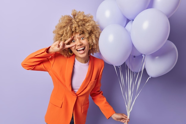 Mooie vrouw met krullend blond haar maakt vredesgebaar over oog gekleed in modieuze oranje jas viert speciale gelegenheid houdt stelletje opgeblazen ballonnen geïsoleerd over paarse achtergrond