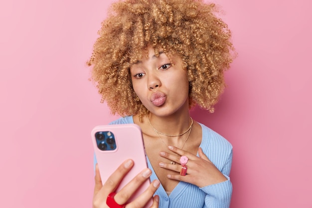 Mooie vrouw met krullend borstelig haar houdt lippen gevouwen maakt selfie poses op smartphone heeft online praten met vriendje spreekt liefde weas casual blauwe trui en sieraden geïsoleerd op roze muur