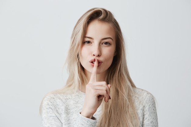 mooie vrouw met lang blond geverfd haar draagt een casual t-shirt met lange mouwen, toont een stilte-teken, heeft een serieuze uitdrukking, vraagt haar haar geheim niet te vertellen, hoopt op loyaliteit en stilte.