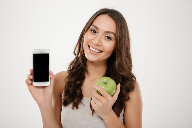 mooie vrouw met perfecte glimlach die zilveren mobiele telefoon op camera aantonen en houden, groene appel die over witte muur wordt geïsoleerd