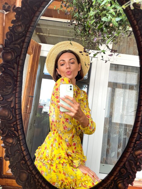 Mooie vrouw thuis maakt foto-selfie in spiegel op mobiele telefoon voor verhalen en berichten op sociale media, met felgele zomerjurk en hoed