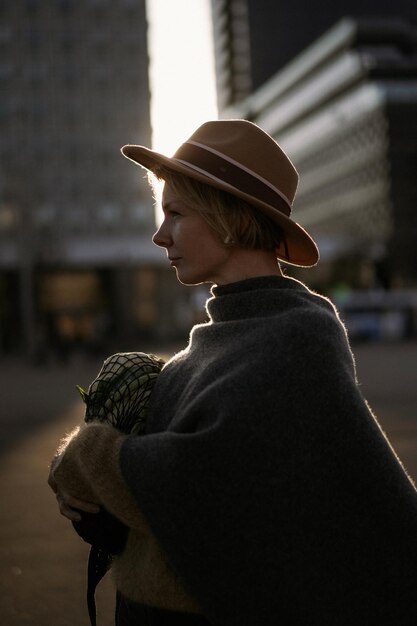 Mooie vrouw van middelbare leeftijd in een hoed met een kort kapsel in het centrum van een grote stad. Close-up portret, zacht tegenlicht.
