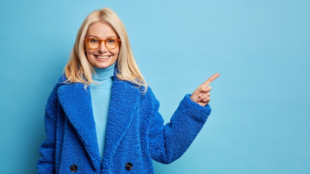 Mooie vrouw van middelbare leeftijd met blond haar draagt een bril en warme blauwe jas wijzend op kopie ruimte