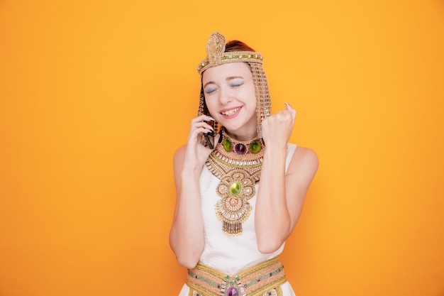 Mooie vrouw zoals cleopatra in oud egyptisch kostuum blij en opgewonden die vuist opheft terwijl ze op mobiele telefoon op oranje praat