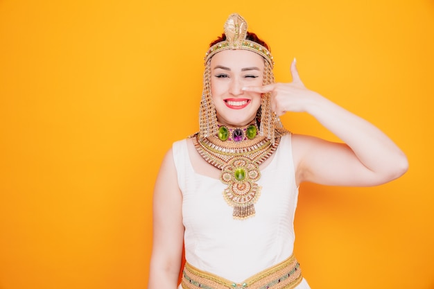 Mooie vrouw zoals cleopatra in oud egyptisch kostuum gelukkig en vrolijk wijzend met wijsvinger naar haar neus glimlachend op oranje