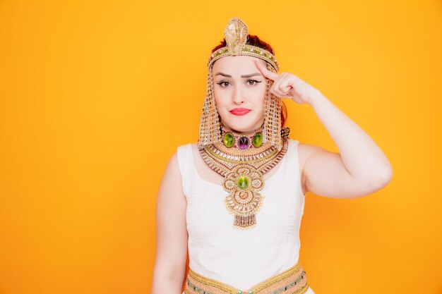 Mooie vrouw zoals cleopatra in oud egyptisch kostuum verward wijzend met wijsvinger naar haar tempel die probeert zich te concentreren op een taak die hard is op oranje