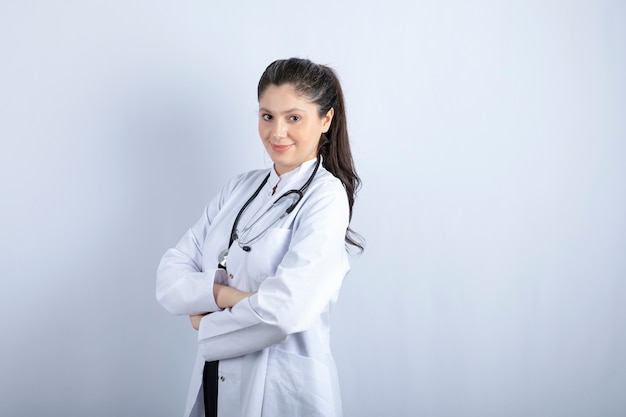 Mooie vrouwelijke arts in witte jas staande armen gekruist over witte muur.