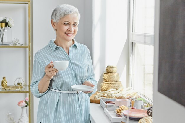 Mooie vrouwelijke gepensioneerde m / v in gestreepte zomerjurk bedrijf schotel en mok, het drinken van thee, poseren in stijlvol interieur met eten en decoraties op de vensterbank. Rijpe vrouw die van ontbijt geniet