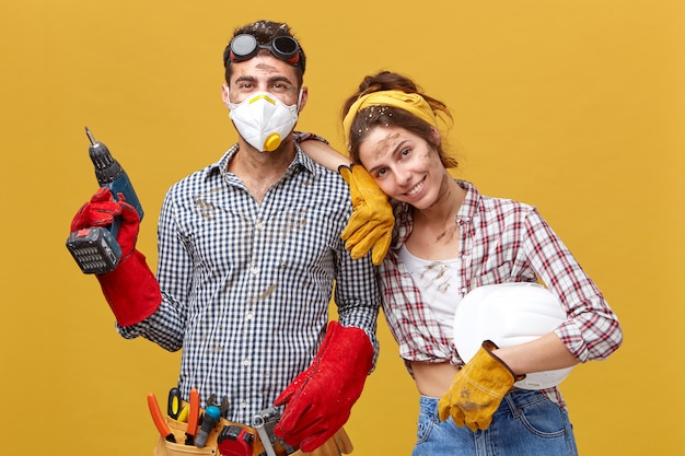 Mooie vrouwelijke huisvrouw die haar echtgenoot helpt met constructie die een geruit overhemd draagt met een witte helm die op zijn schouder leunt met een goed humeur en blij is om hem te helpen. Mensen en relaties