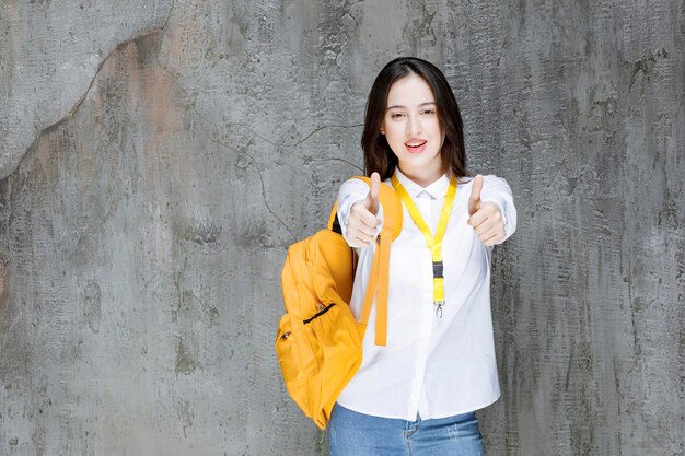 Mooie vrouwelijke student met gele rugzak duimen opgevend. Hoge kwaliteit foto