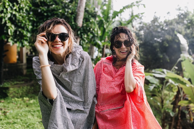 Mooie vrouwen met donker nat haar die tijdens wandeling lachen. Buitenfoto van prachtige dames in regenjas die genieten van trekking.