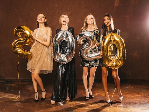 Mooie vrouwen Nieuwjaar vieren. Gelukkige prachtige meisjes in stijlvolle sexy feestjurken met gouden en zilveren 2020-ballonnen, plezier op oudejaarsfeest. Vakantieviering. Charmante modellen