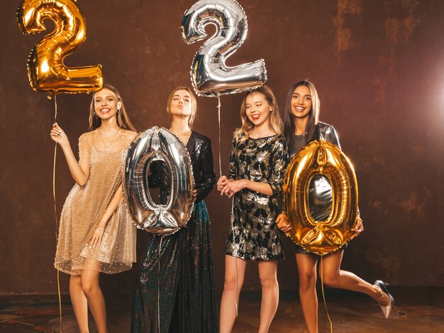 Mooie vrouwen Nieuwjaar vieren. Gelukkige prachtige meisjes in stijlvolle sexy feestjurken met gouden en zilveren 2020-ballonnen, plezier op oudejaarsfeest. Vakantieviering. Charmante modellen