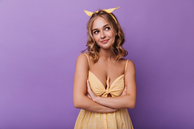 Mooie witte vrouw in trendy zomerjurk poseren met gevouwen armen. Indoor portret van geïnteresseerde charmante dame met golvend haar geïsoleerd op violet.