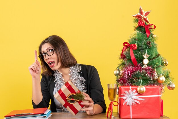 Mooie zakelijke dame in pak met bril die verrassend omhoog wijst en zittend aan een tafel met een kerstboom erop in het kantoor op geel