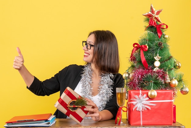 Mooie zakelijke dame in pak met bril ok gebaar zittend aan een tafel met een kerstboom erop in het kantoor