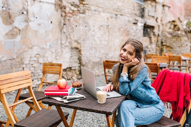 Mooie zakenvrouw met lang blond haar witte laptopcomputer gebruikt tijdens de lunchpauze op terras op bakstenen muur achtergrond. mooi meisje dat spijkerbroek draagt, zittend aan de houten tafel.