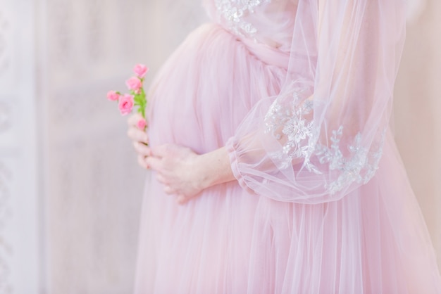 Mooie zwangere vrouw in rijk roze jurk houdt handen op haar buik poseren