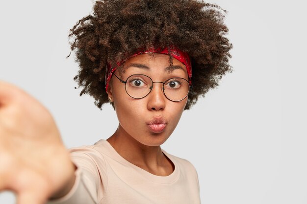 Mooie zwarte oman pruilt lippen terwijl selfie de handen naar voren strekt, een transparante bril draagt, een casual trui