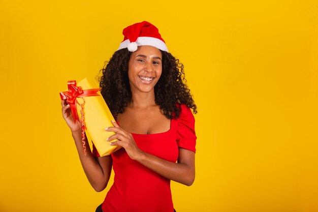 Mooie zwarte vrouw in kerstman hoed met vrolijk kerstcadeau op gele achtergrond.